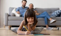Petite fille couchée sur le sol en train de dessiner avec ses parents assis dans le divan derrière elle